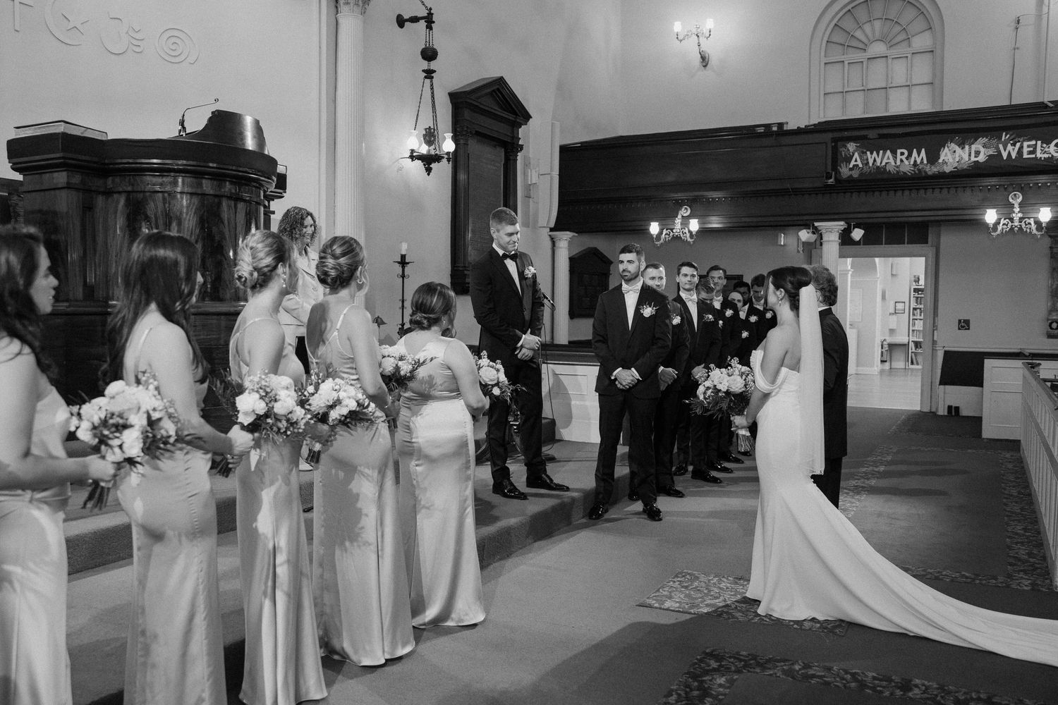 Wedding party members in formal attire stand in line at an indoor venue.
