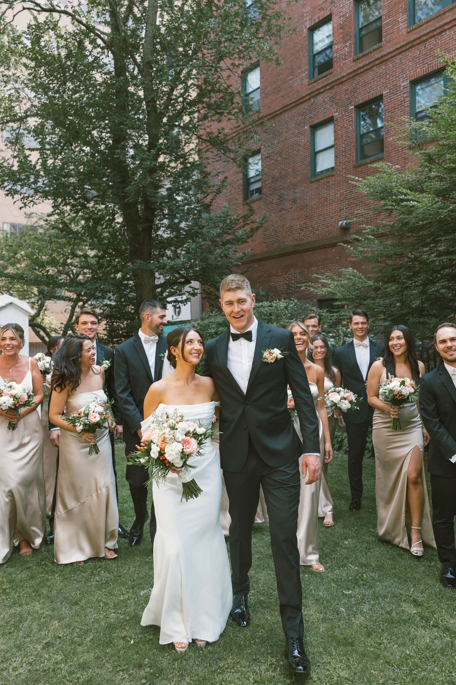 Wedding party poses on grass outside brick building wearing formal attire.