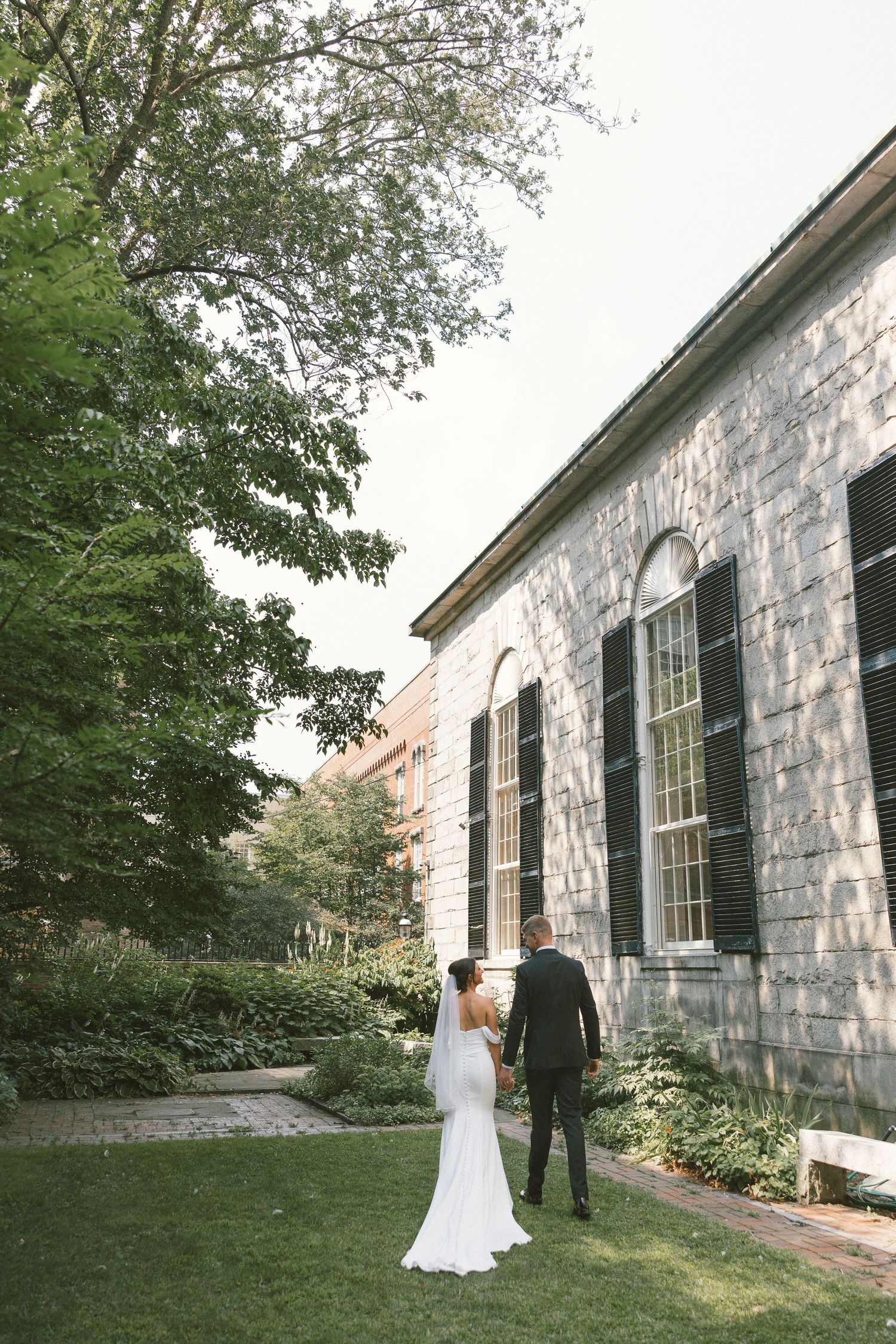 Wedding couple poses for photos against stone building exterior with green landscaping.