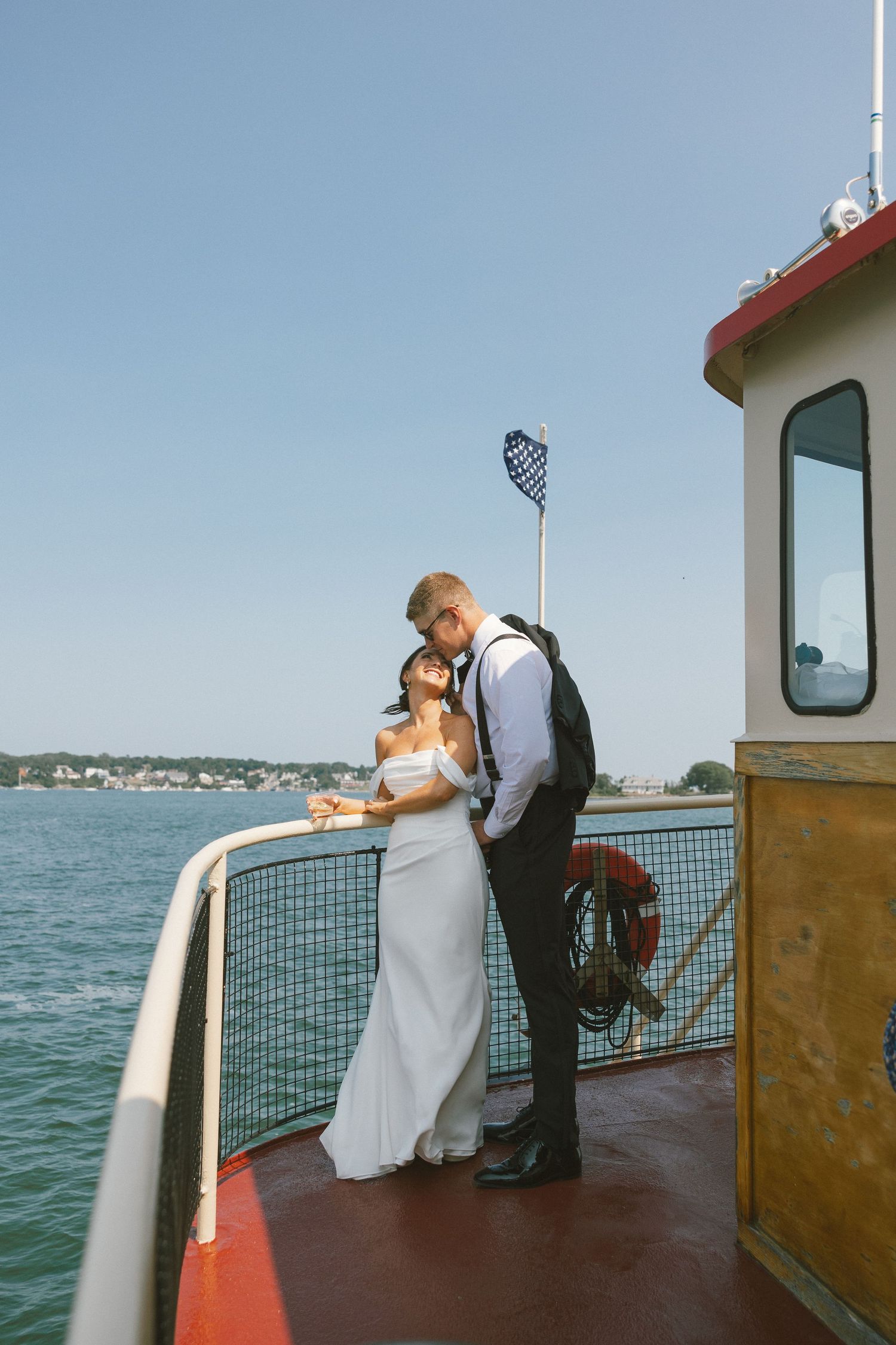 Wedding couple poses on deck of boat with ocean and coastline in background.