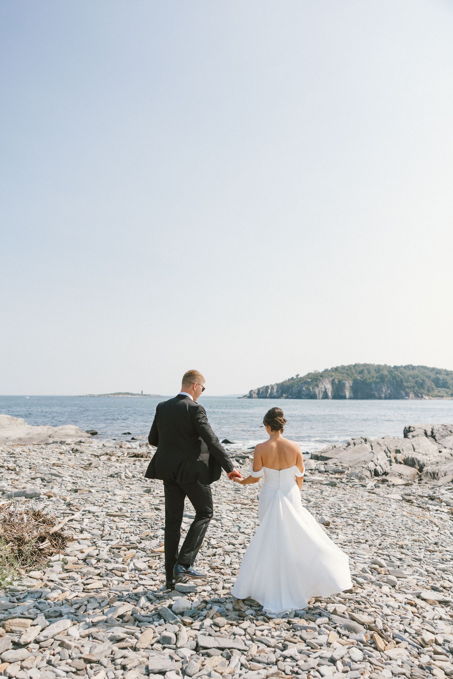 Wedding couple walks along rocky beach shore with ocean and islands in background.