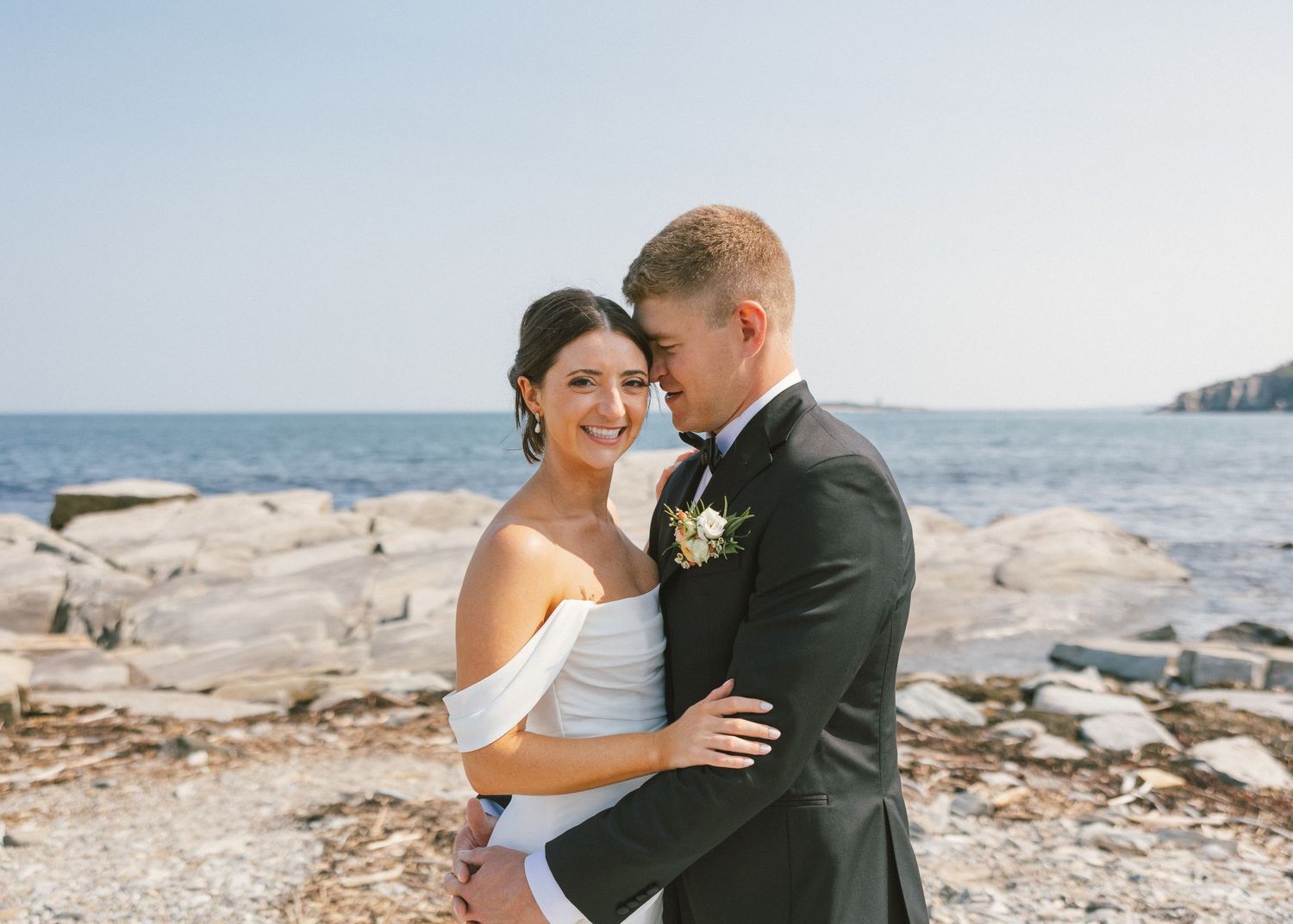 Romantic coastal wedding photoshoot on a rocky beach with ocean backdrop.