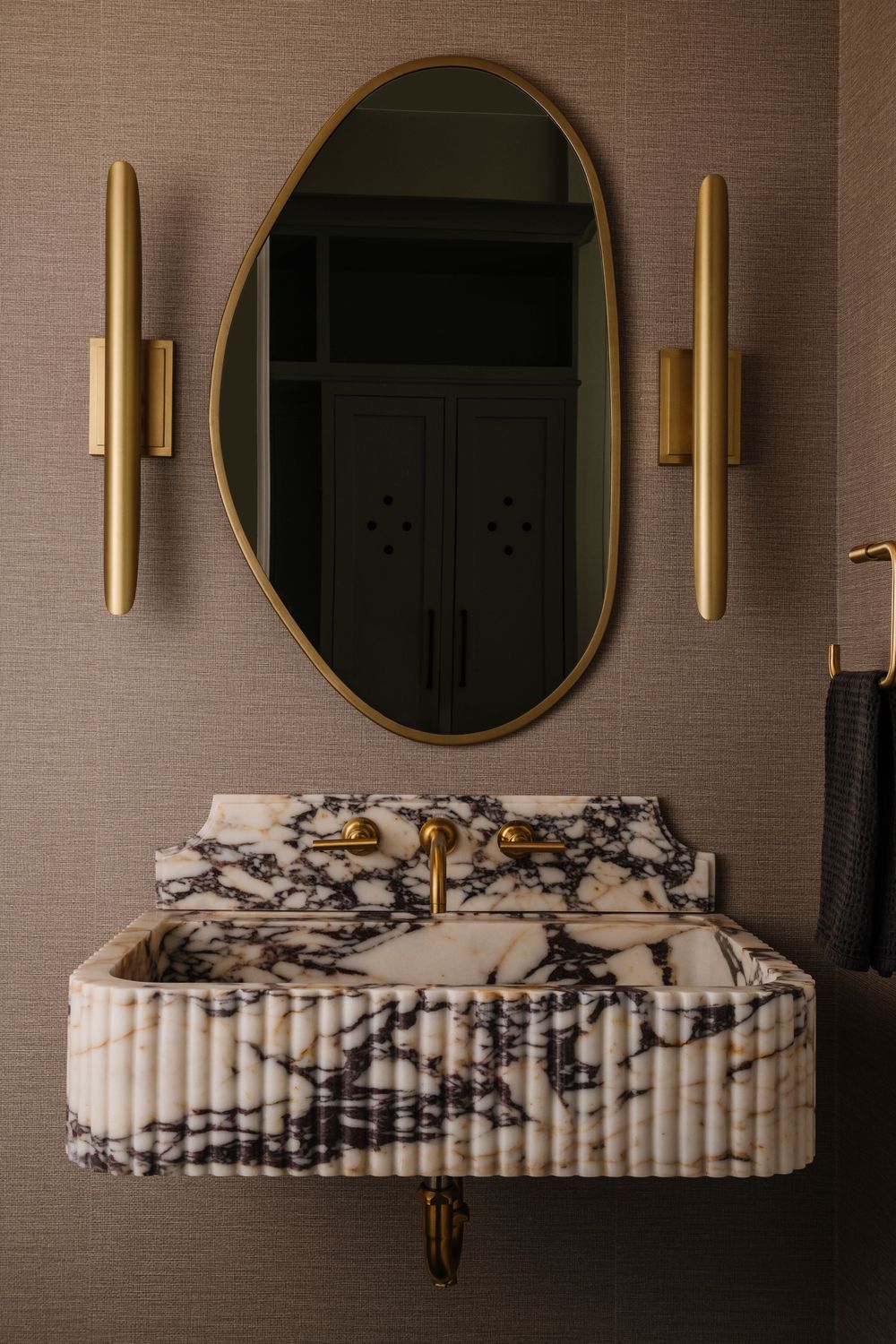Marble sink in powder bath with dark wallpaper and brass sconces.