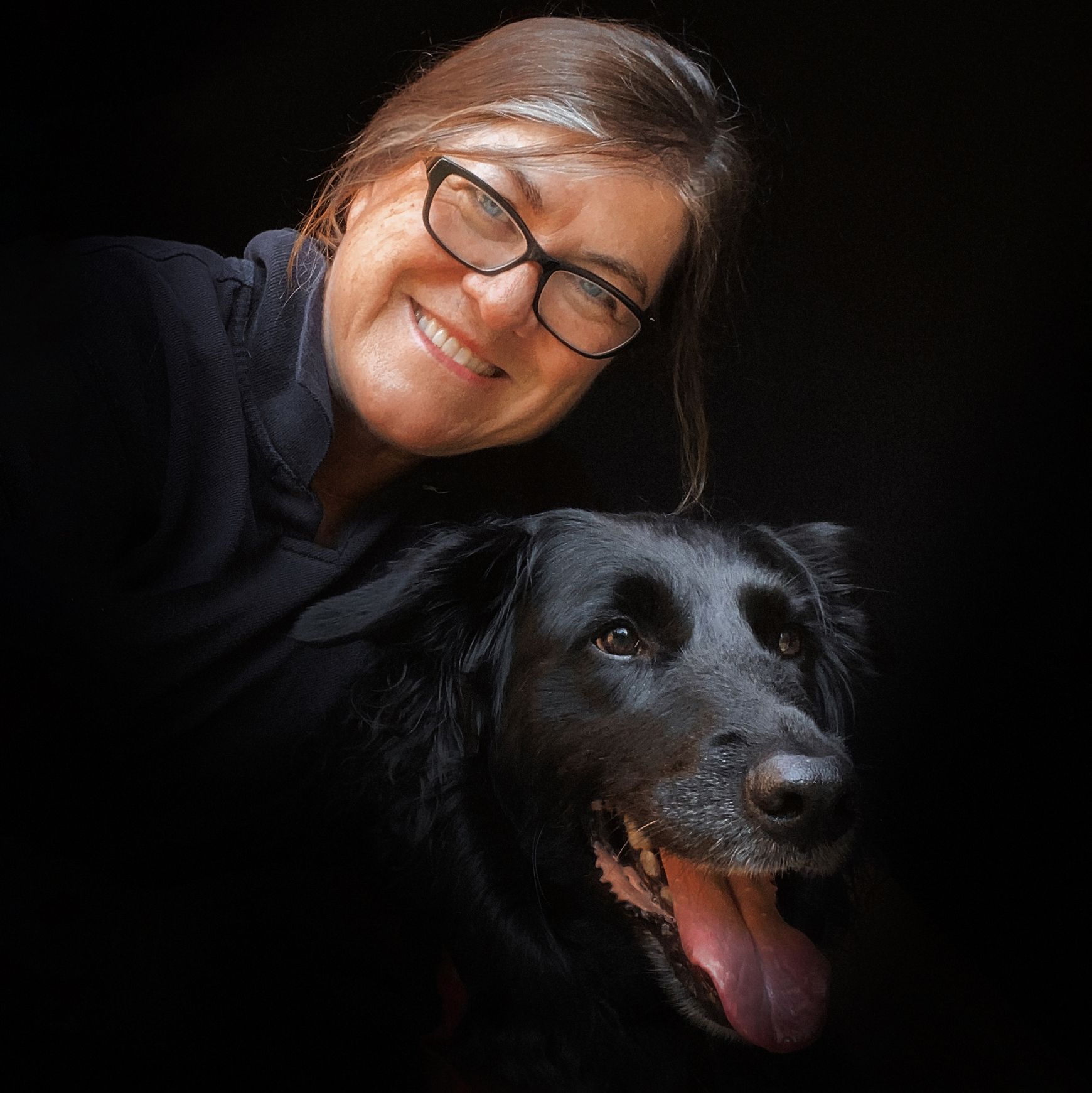 A black dog with a happy expression poses with someone in glasses against a dark background.