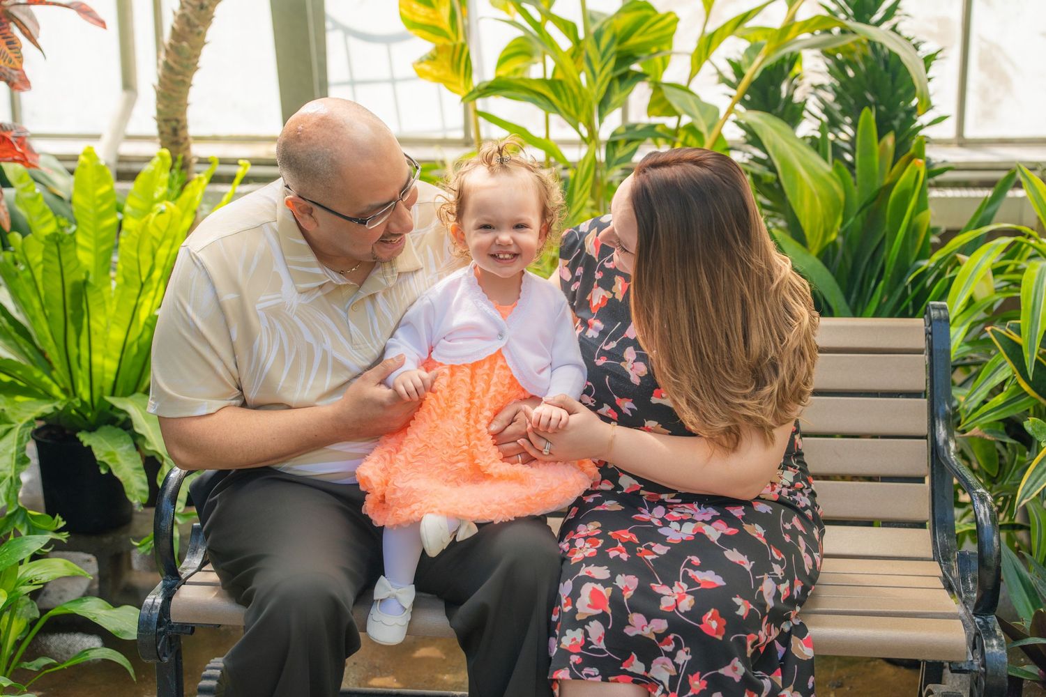 A family of three sits on a bench in a lush greenhouse, parents smiling at toddler daughter in an orange dress. Quad Cities Photography