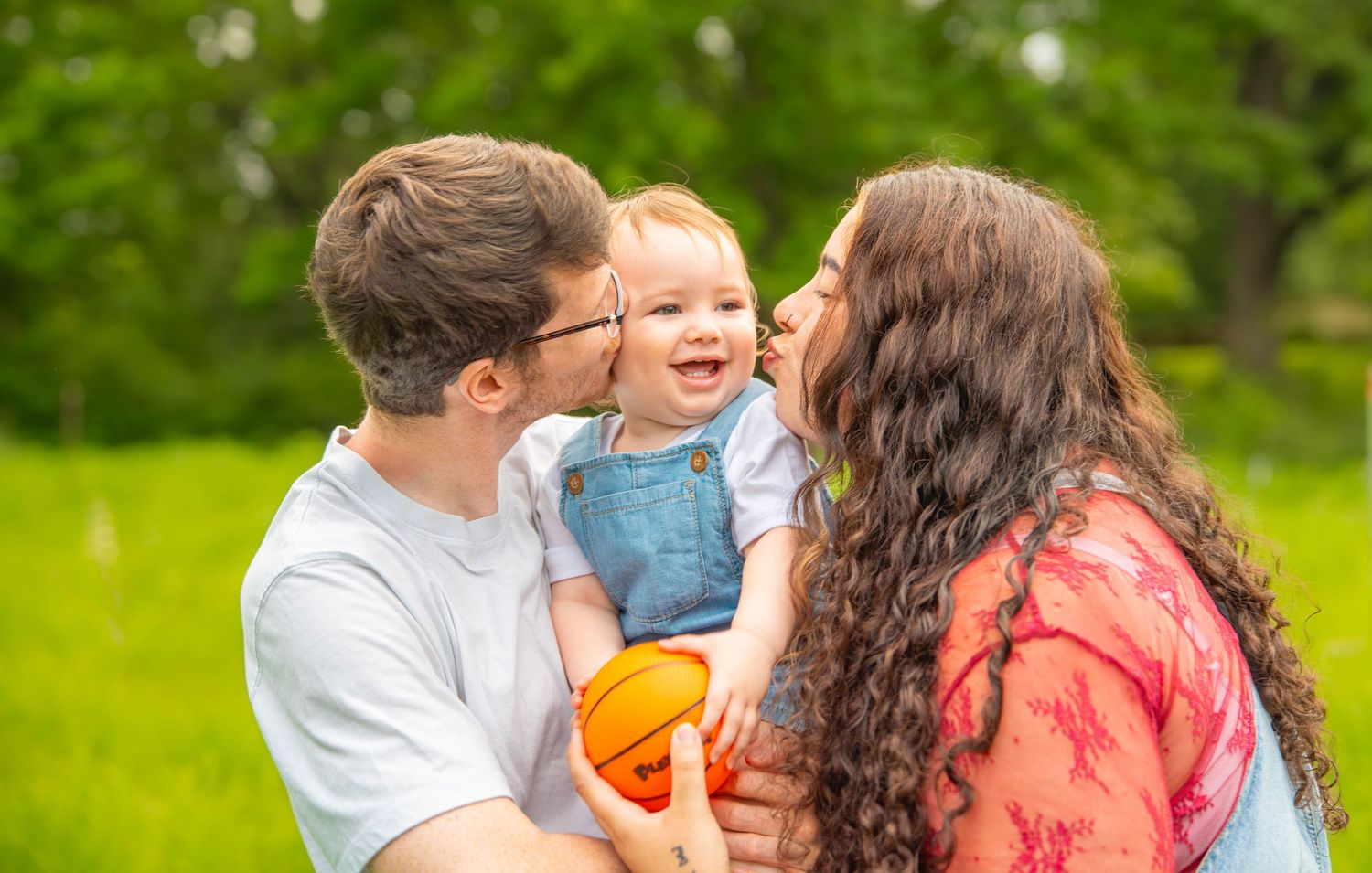Happy family with parents kissing smiling baby outdoors in lush green park setting. Quad Cities Photography