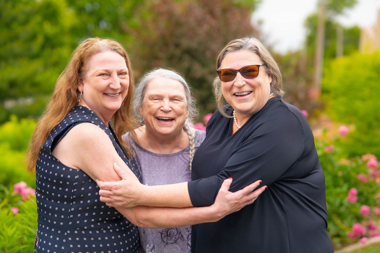 Three women smiling and embracing outdoors in a lush green garden with pink flowers blooming in the background.