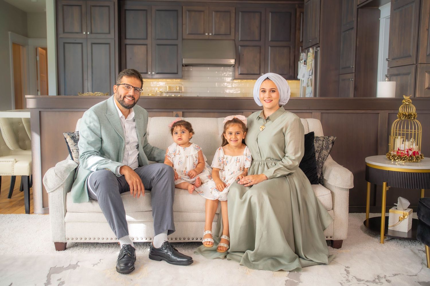 A family sitting together on a grey couch in an elegant home interior with wooden cabinetry and neutral decor.