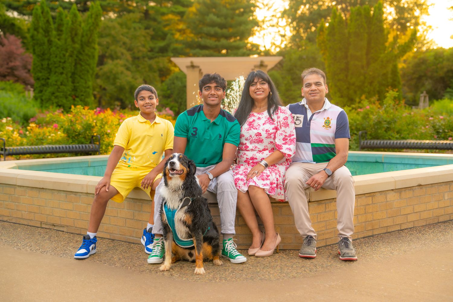 A group sits on a fountain ledge with their Cavalier King Charles Spaniel dog during golden hour in a garden setting.