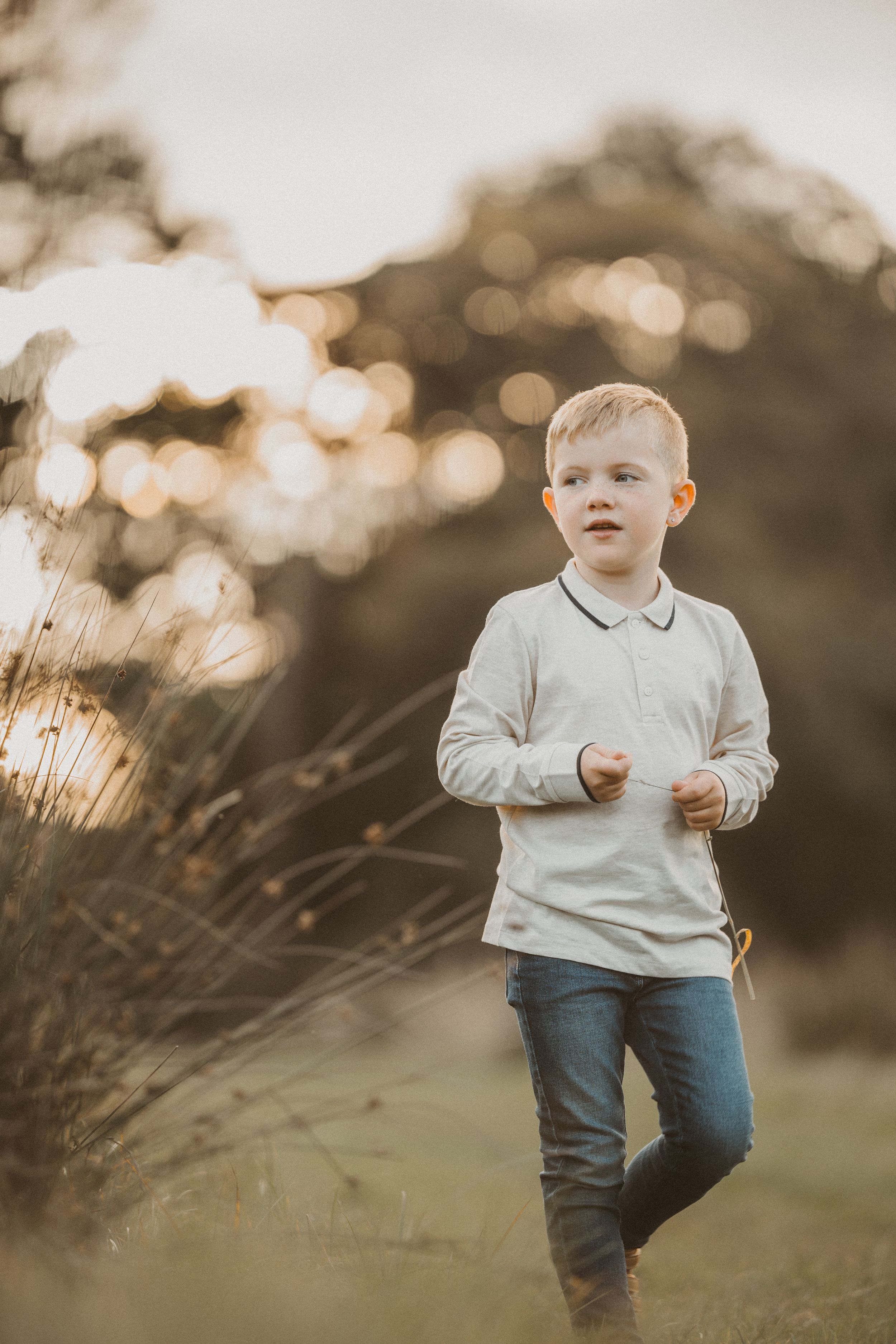 A toddler in a white sweater and jeans explores an outdoor field during golden hour sunset.