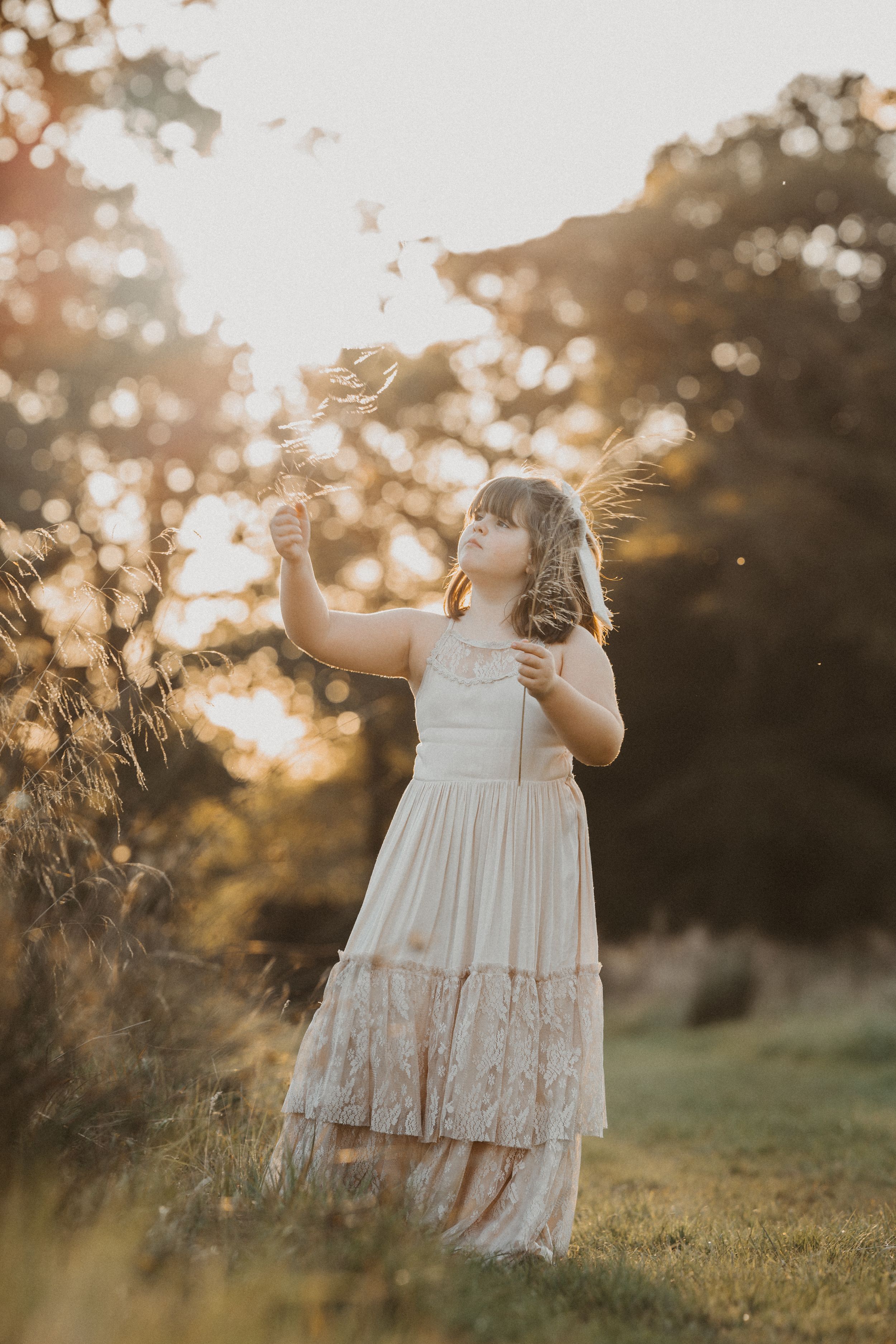 A child in a white lace dress stands in a golden sunlit field reaching up toward falling flower petals at sunset.