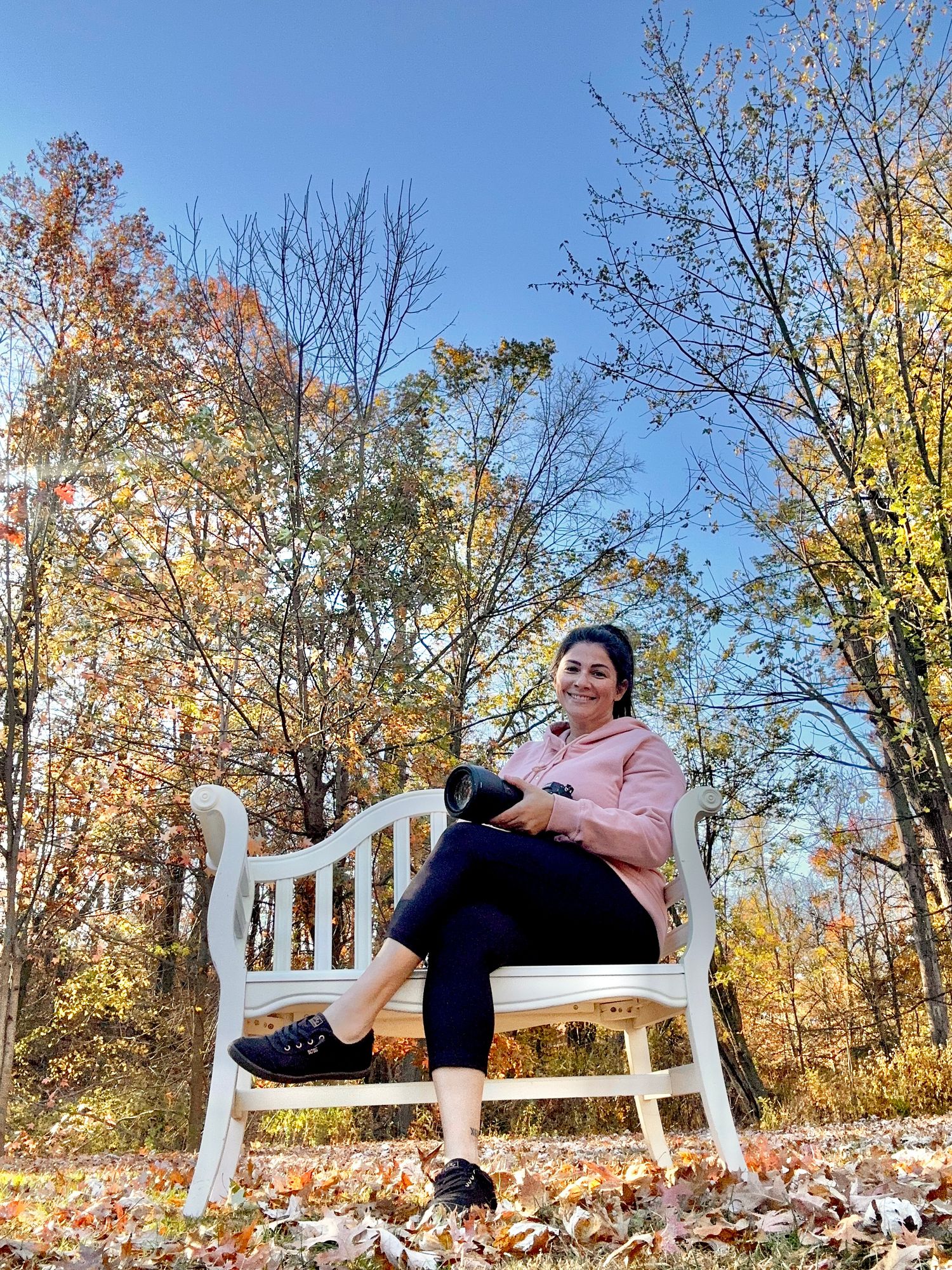 A person sits on a white wooden bench surrounded by colorful autumn foliage and fallen leaves on a sunny day.