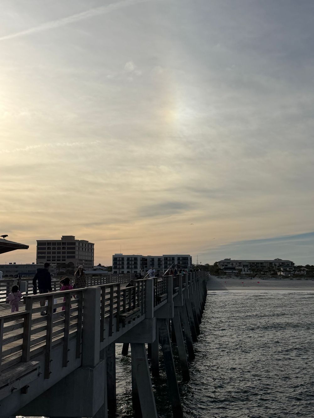 Wooden pier extends over calm waters at dusk with buildings visible along the shoreline.