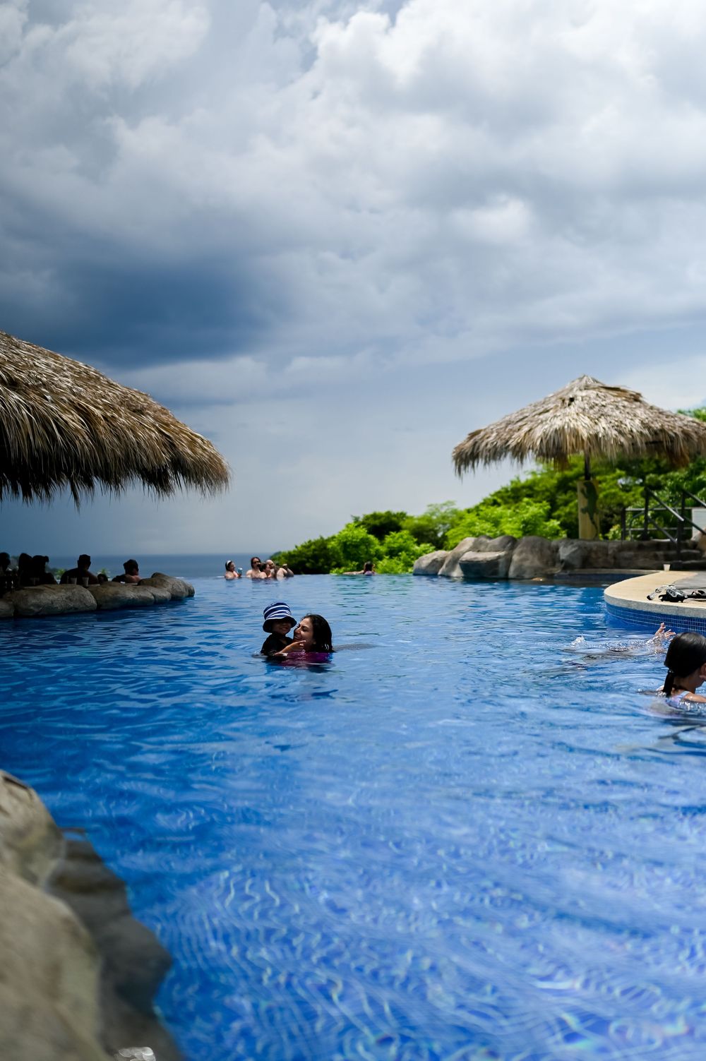 An infinity pool overlooking the ocean with thatched umbrellas and cloudy skies.