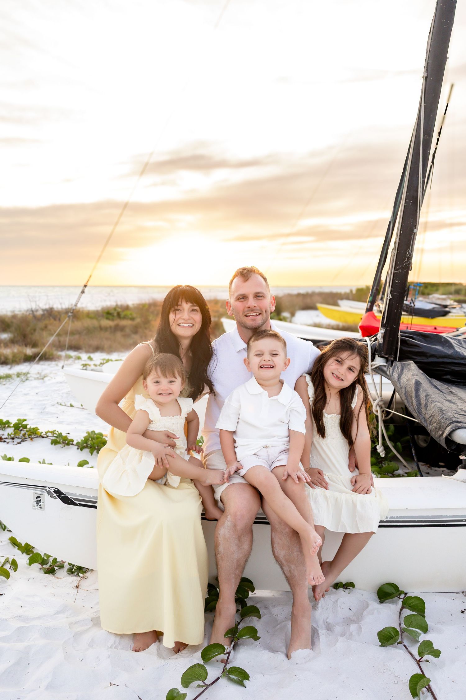 Happy family of four with two young children pose outdoors on a sunny tree-lined gravel path.