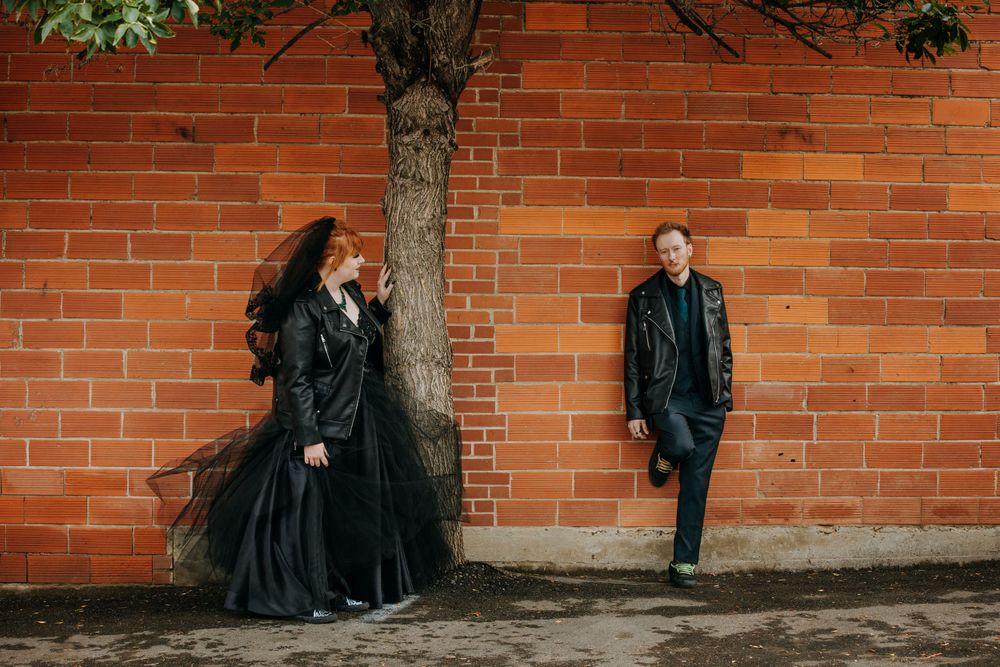 Alternative couple in black attire poses against orange brick wall with tree.