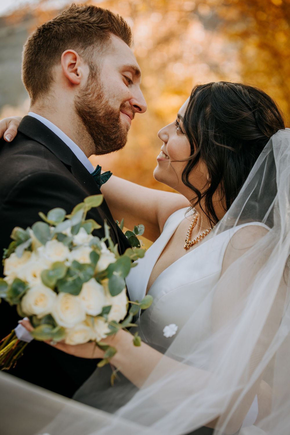 Tender wedding moment with white roses and flowing veil in autumn setting.