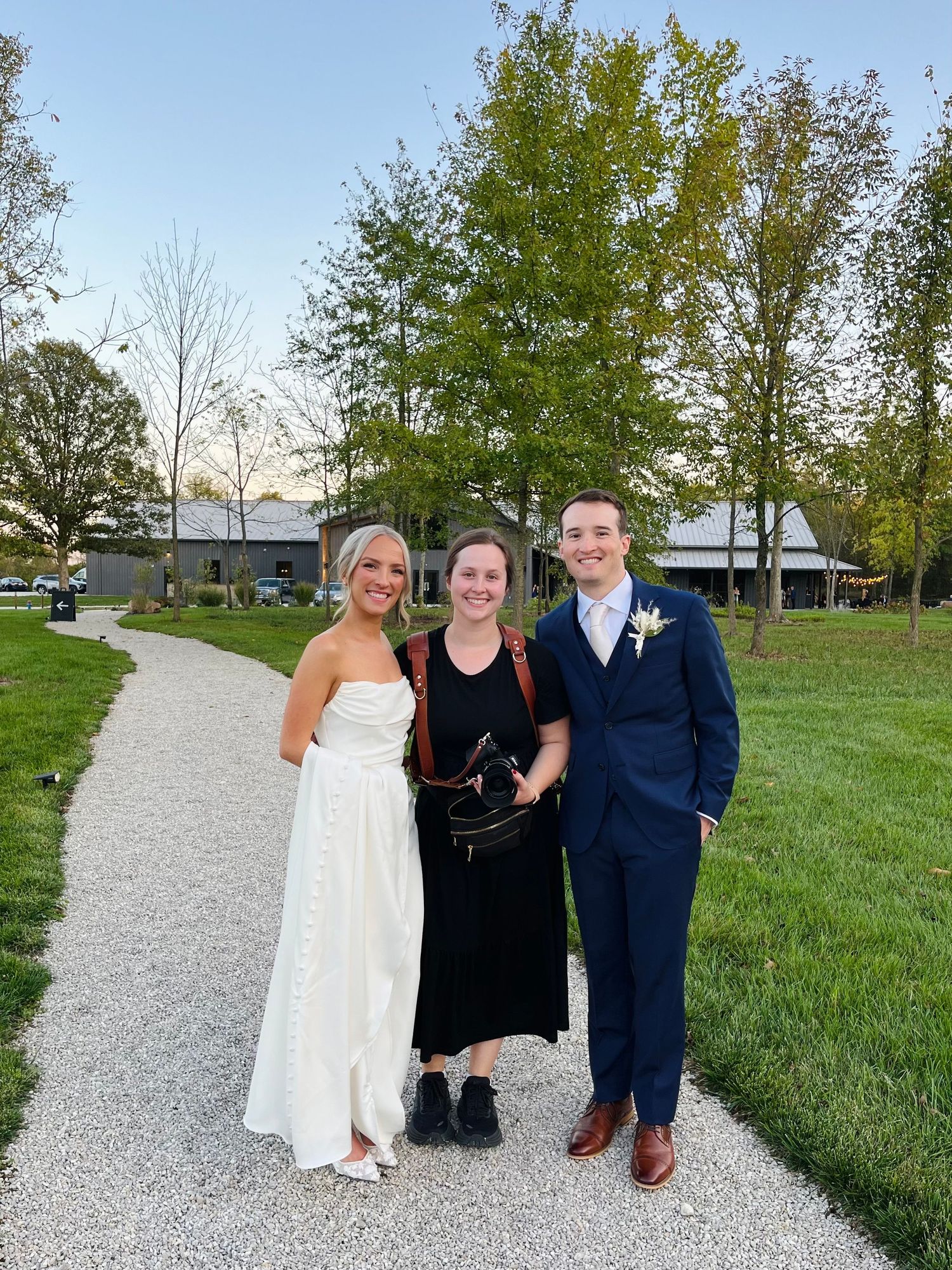 Wedding photographer poses with couple on white gravel path surrounded by green lawn.