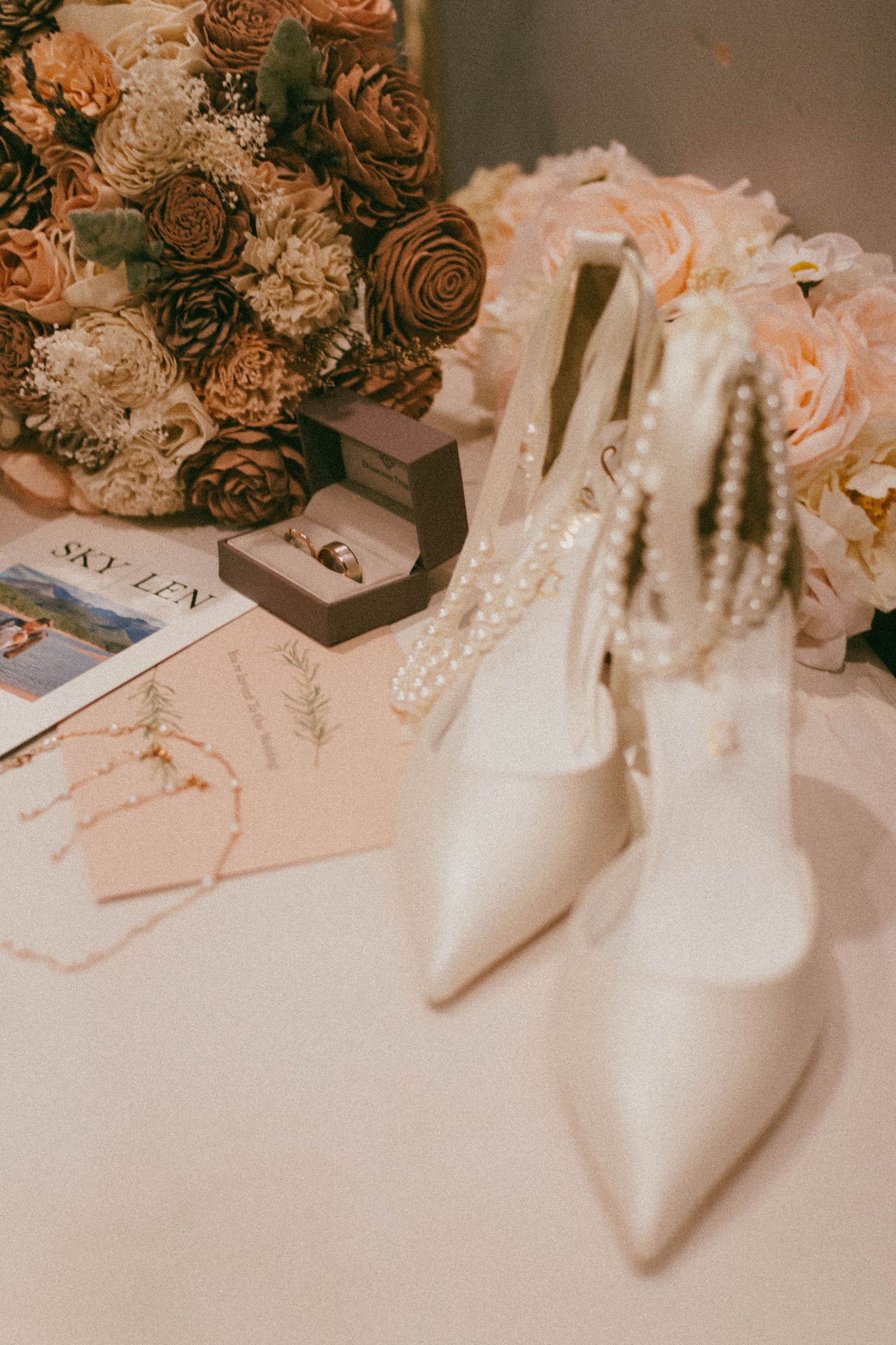 White bridal shoes and wedding details with peach and brown roses arranged with ring box on light surface.