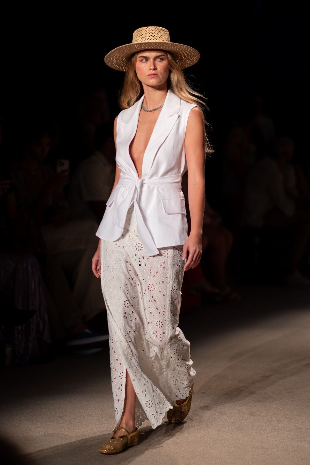Model walking runway in white blazer, lace skirt and straw hat during fashion show.