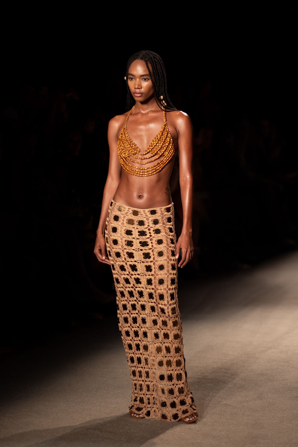Model on catwalk wearing golden beaded top and crochet maxi skirt with geometric pattern.