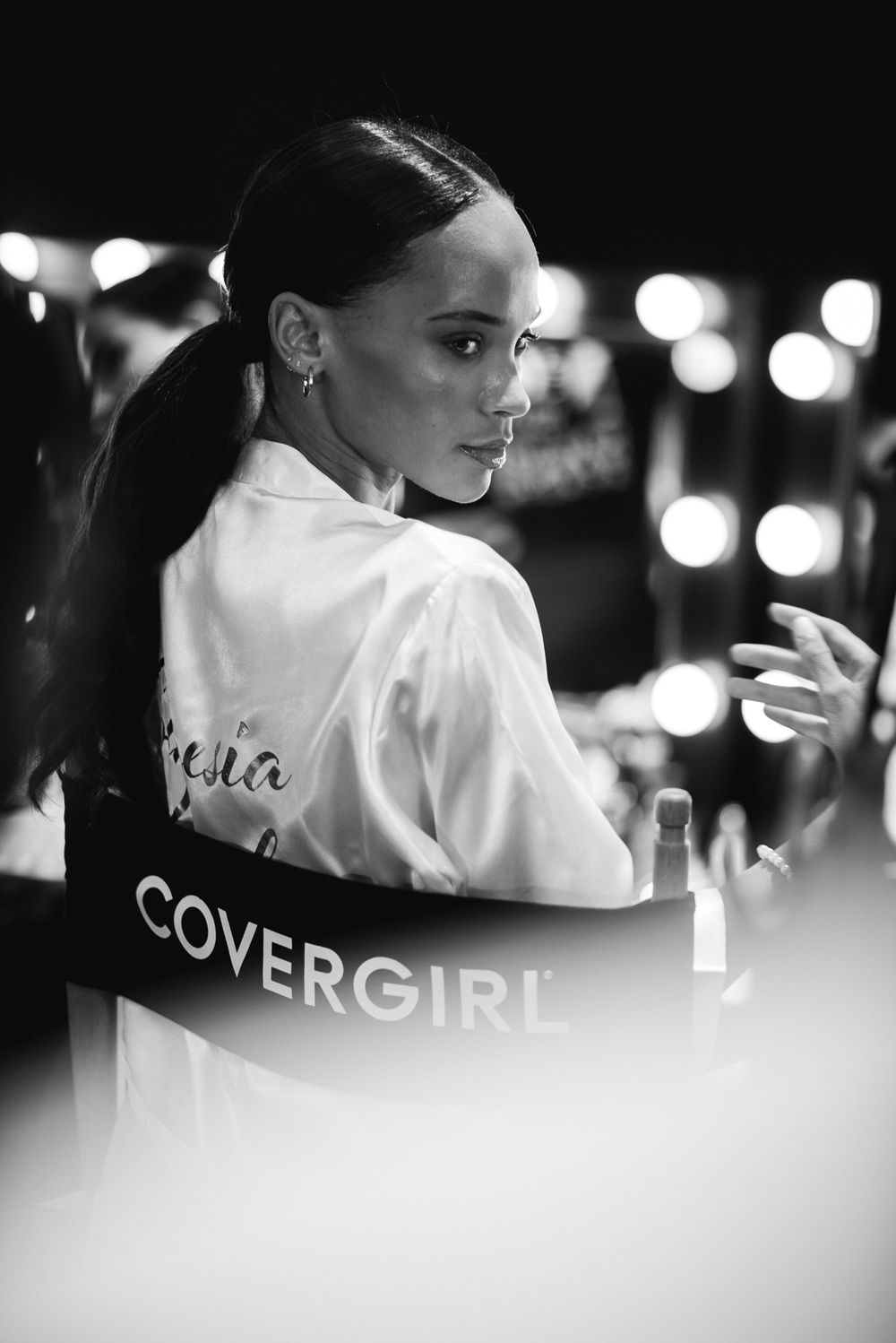 Black and white behind the scenes image of a model wearing a white robe with Covergirl branding at a photoshoot.