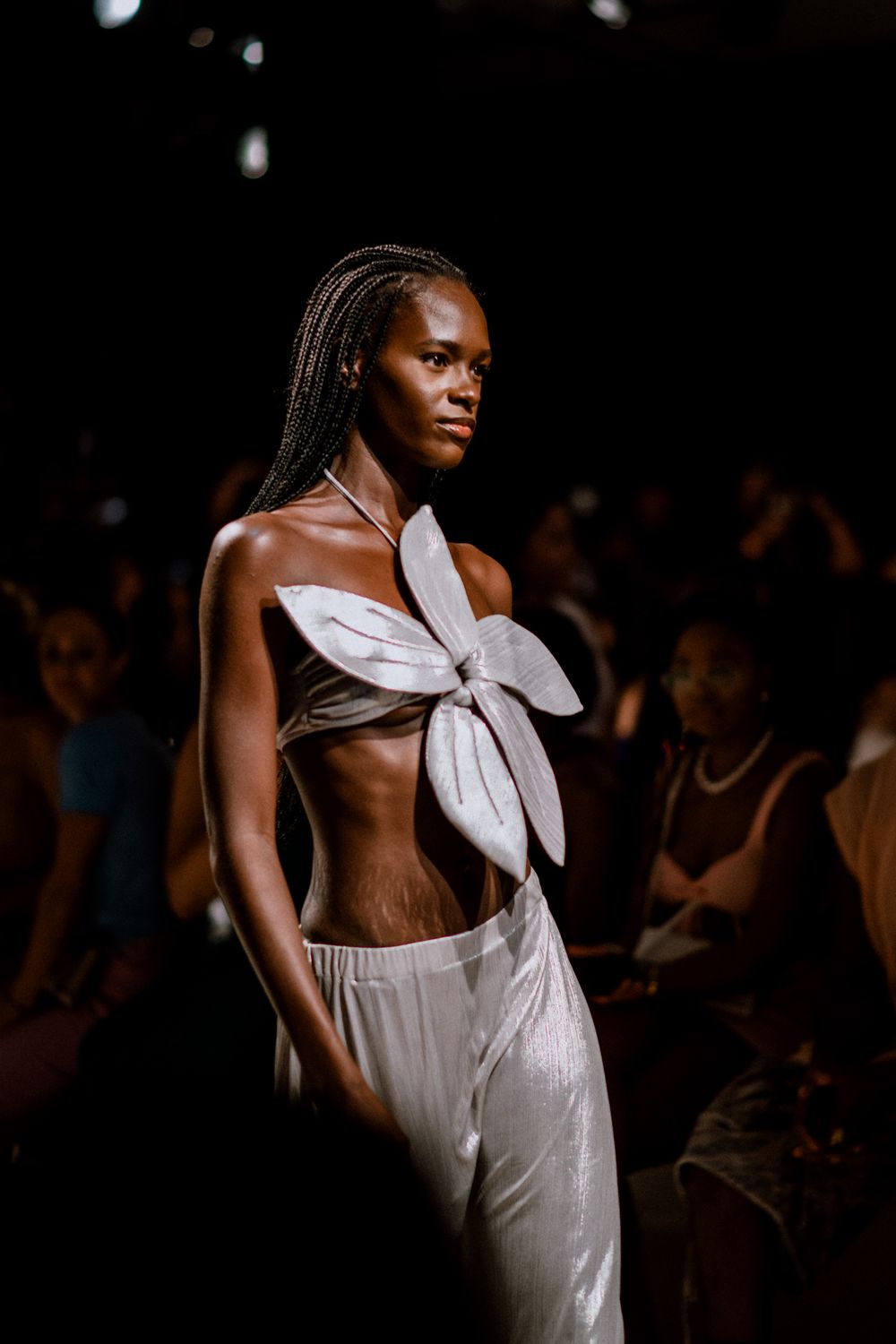 Model walking runway in white bow crop top and light pants during fashion show against dark background.