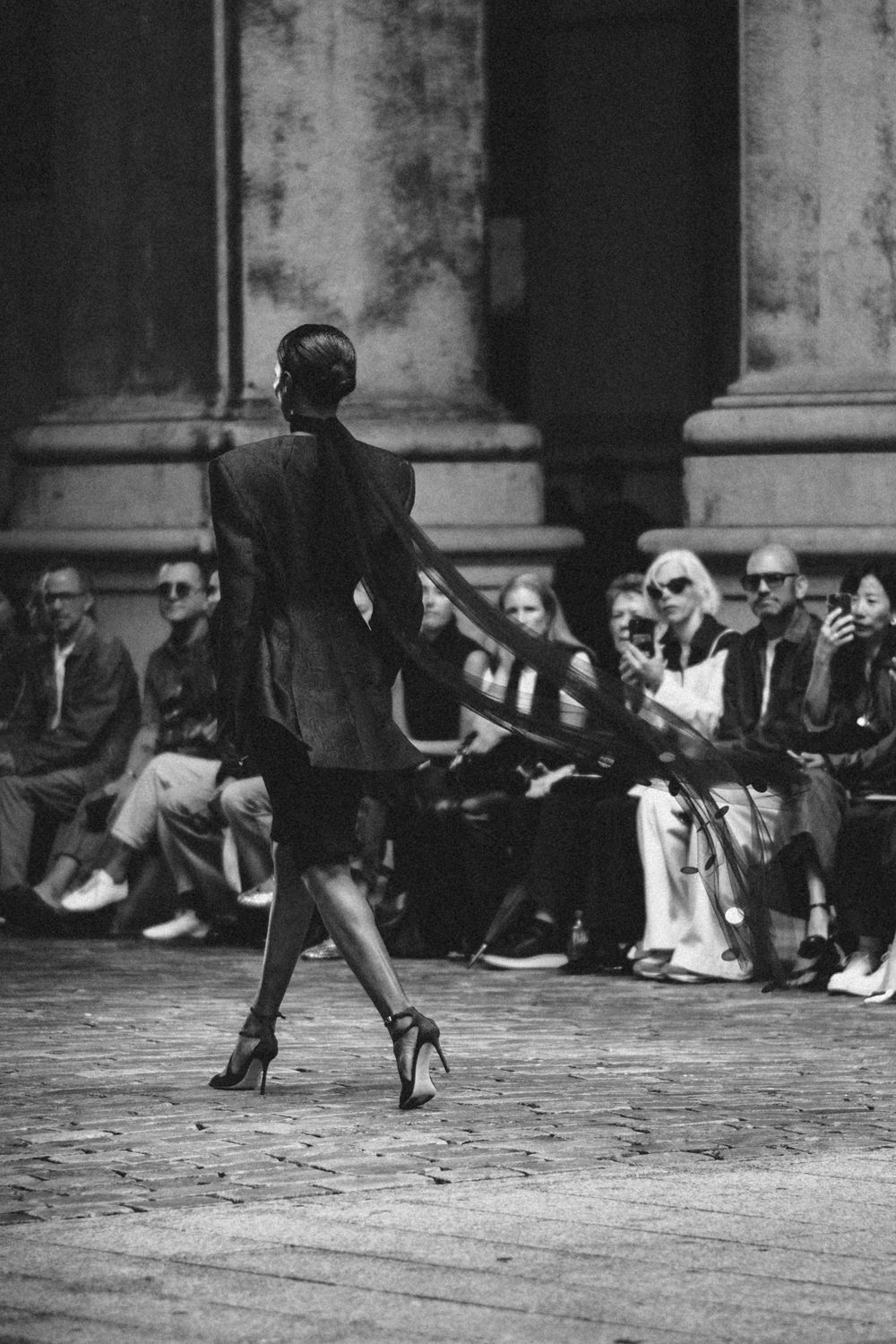 A model walks down the runway in a black outfit and high heels as spectators watch from the sidelines during a fashion show.
