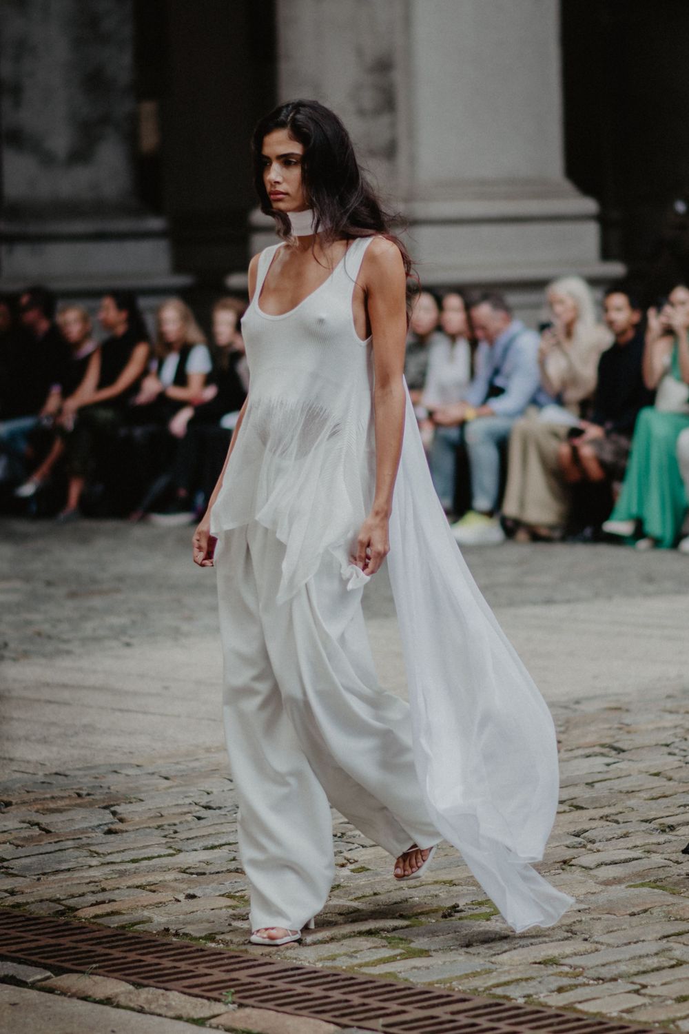 Model walks runway in flowing white ethereal dress with wide-leg pants during outdoor fashion show on cobblestone street.