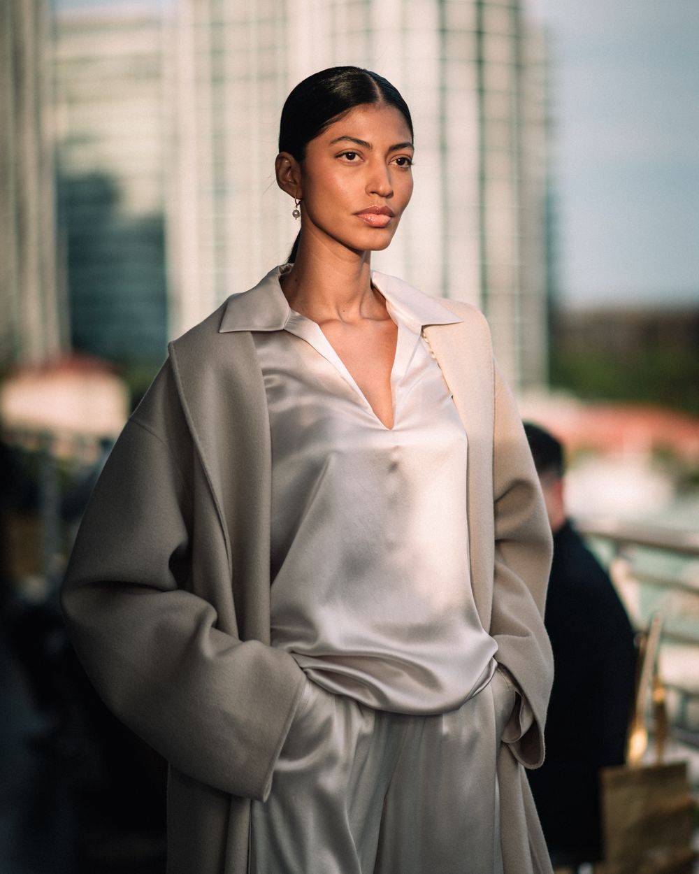 A woman in an oversized beige coat and white blouse poses against a blurred urban cityscape background.