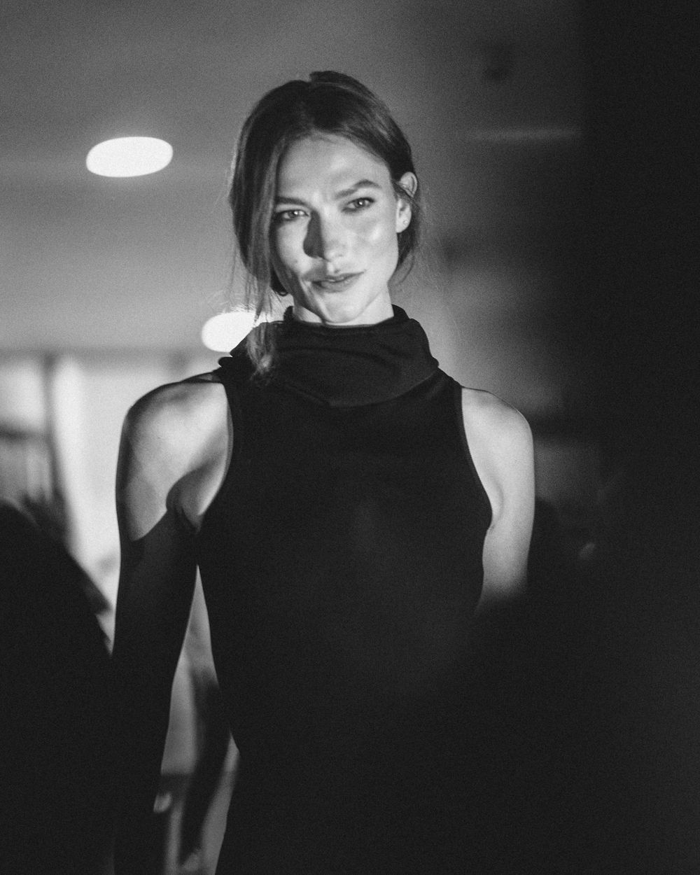 Black and white portrait of a model in a sleeveless turtleneck top with dramatic lighting and blurred background.