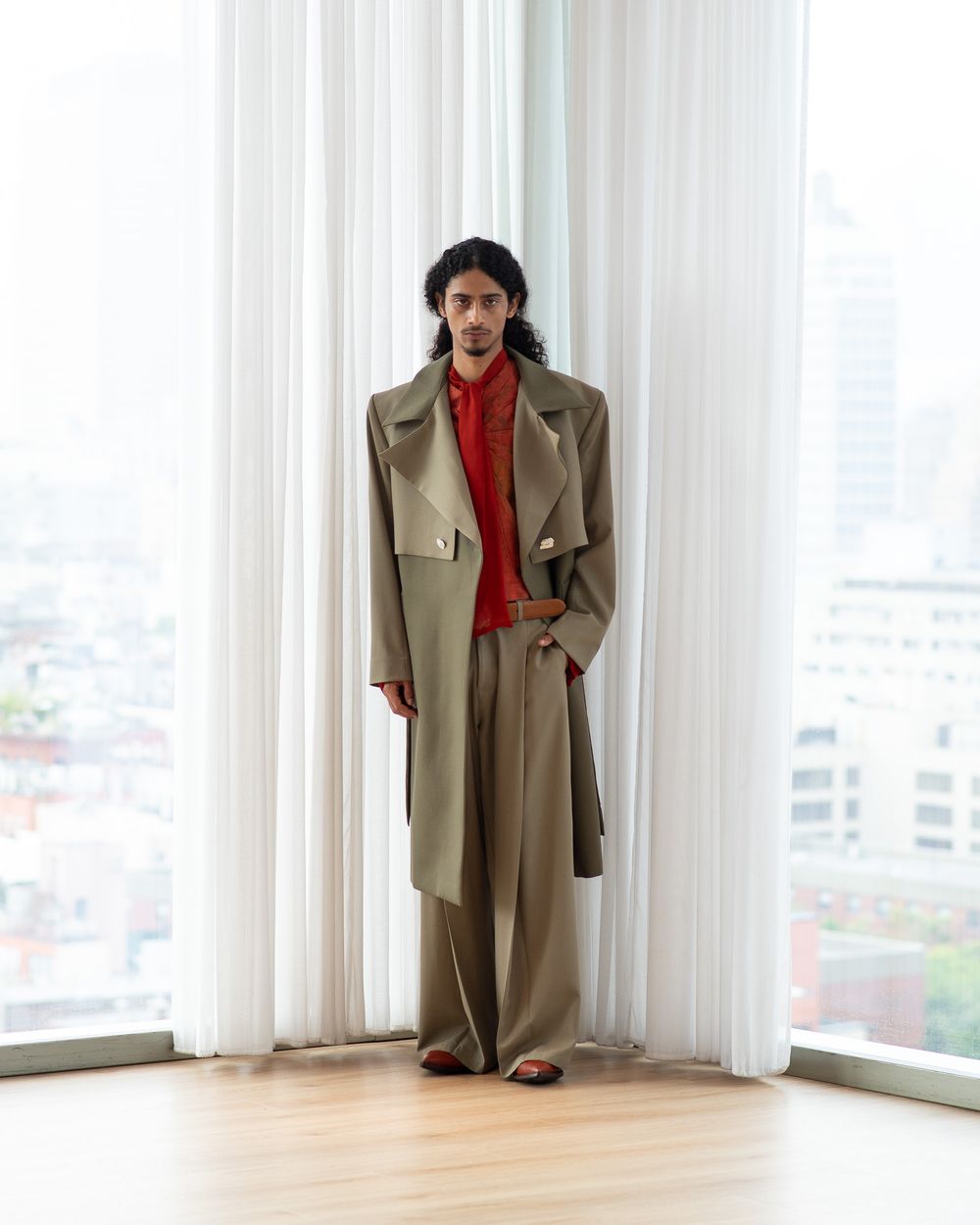 Fashion model wearing oversized beige trench coat and red shirt poses against white curtains in high-rise apartment.