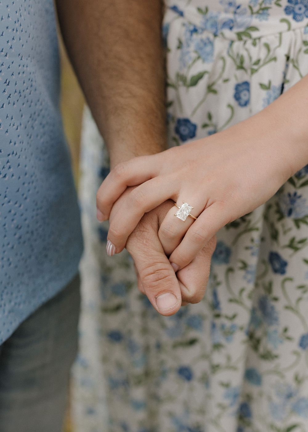 Close-up view of two hands holding each other with a sparkling diamond engagement ring against a floral print dress.