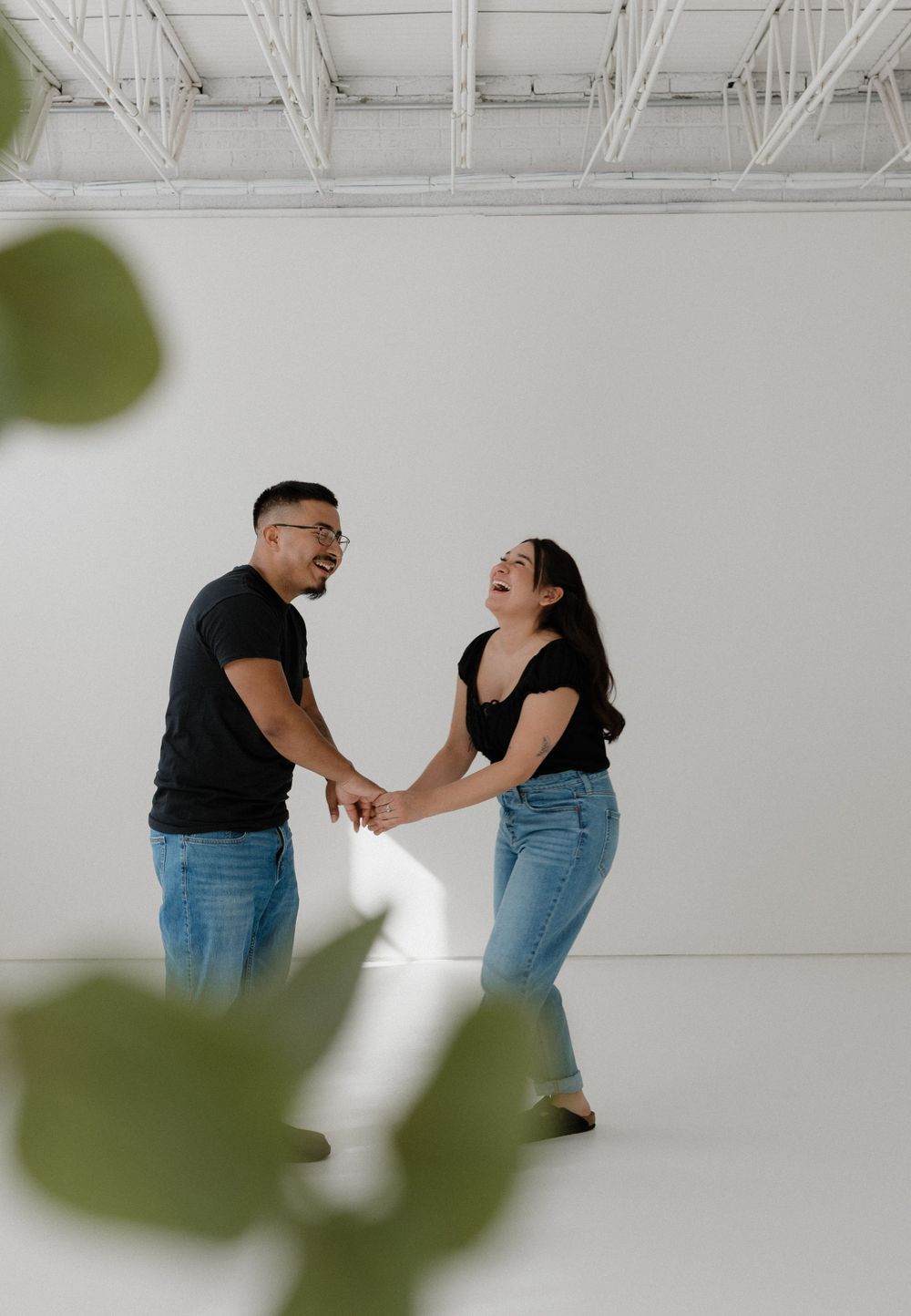 Two people in matching blue jeans and dark tops laugh joyfully together against a white studio backdrop with green leaves in frame.