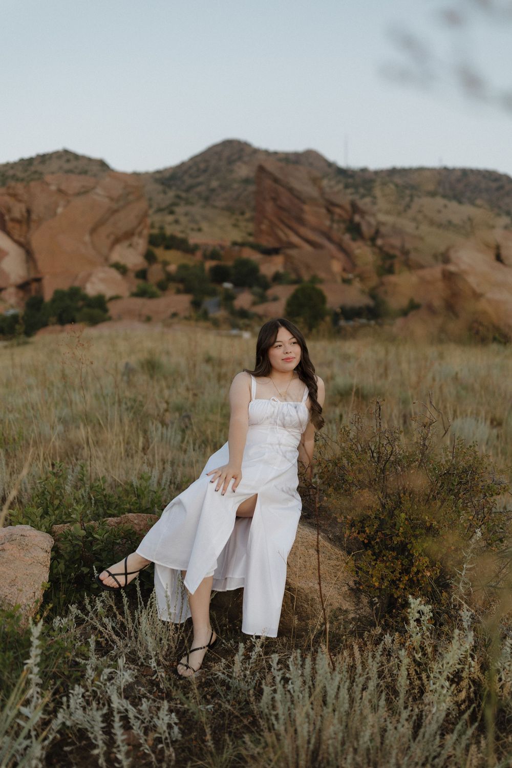 Person in white dress poses against dramatic red rock formation and mountains in outdoor landscape setting.