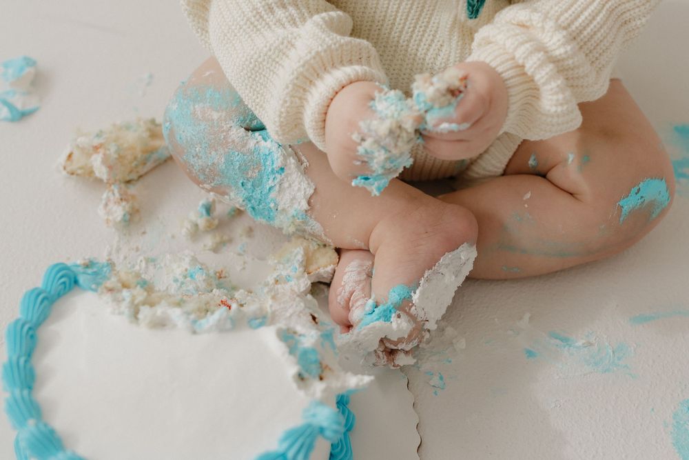 Small hands play with crumbled cake and blue frosting during a festive cake smash photo session.