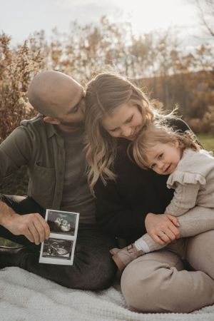 A family shares a tender moment on a blanket outdoors while looking at ultrasound photos during golden hour.