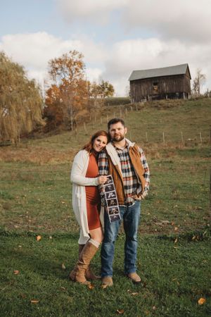 Couple in fall clothing posing together on grassy hillside with rustic barn in background.