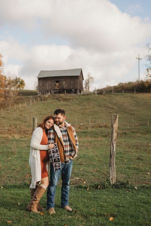 Couple standing close together on autumn hillside with weathered barn above them.
