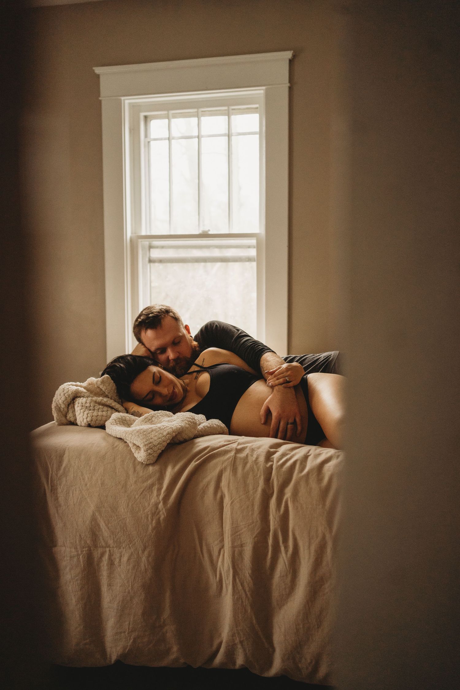 Couple resting together on bed by window in moody natural lighting.