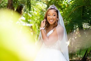 Bride in white dress and tiara poses outdoors with natural greenery backdrop.