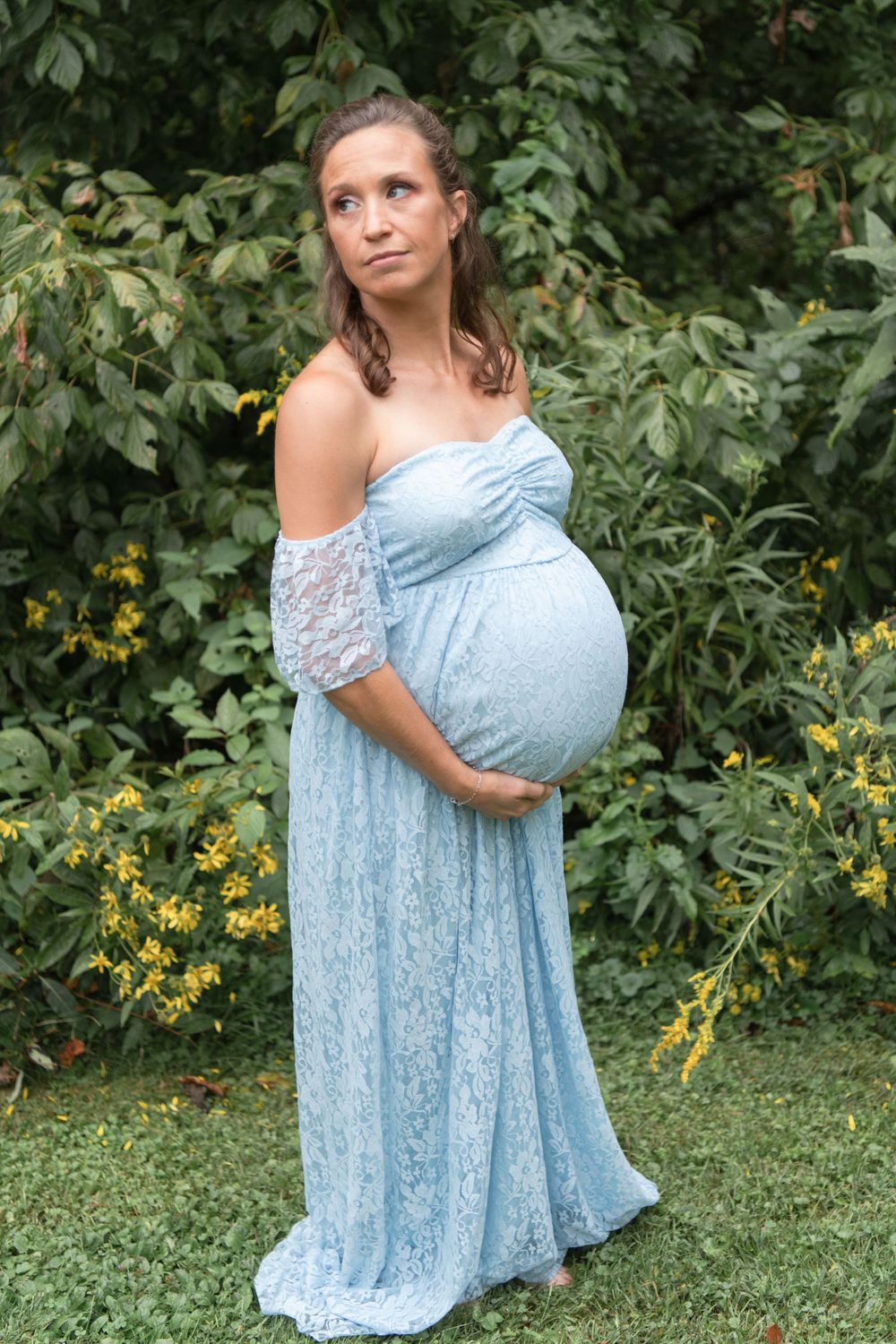 Pregnant woman in blue lace dress stands in garden setting with yellow flowers and greenery.