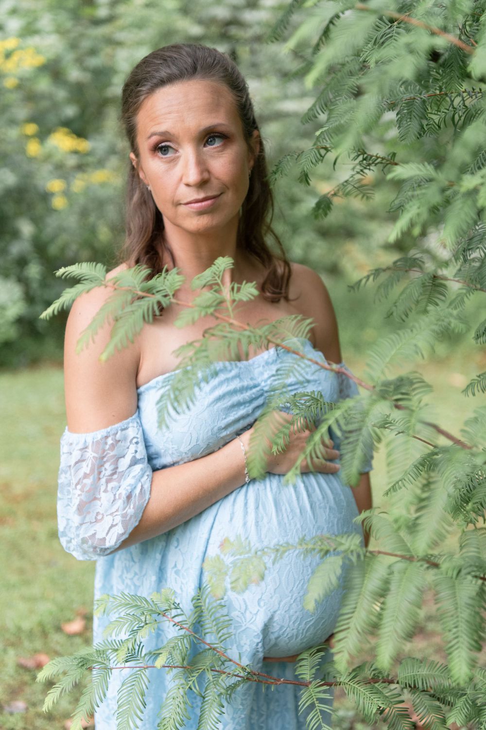 Maternity portrait through fern leaves showing woman in pale blue lace dress outdoors.