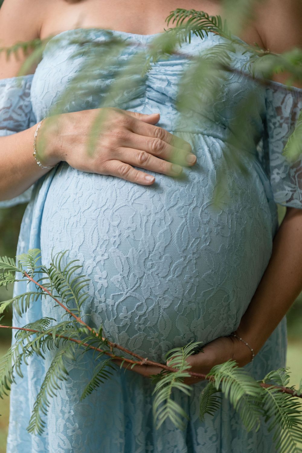 Close-up of pregnant belly in blue lace dress framed by delicate fern leaves.