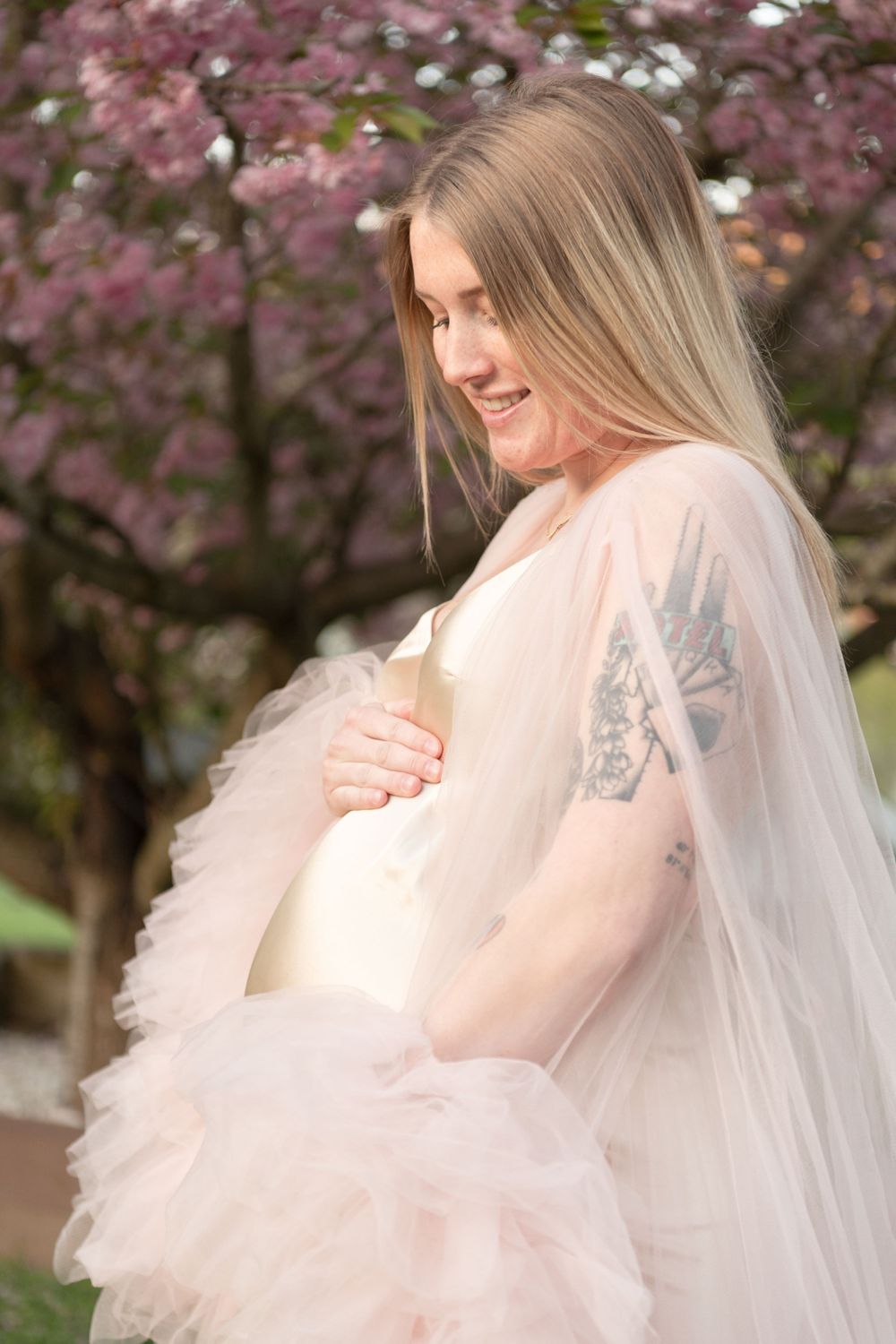 A maternity photo under cherry blossoms wearing a flowing pink tulle dress.