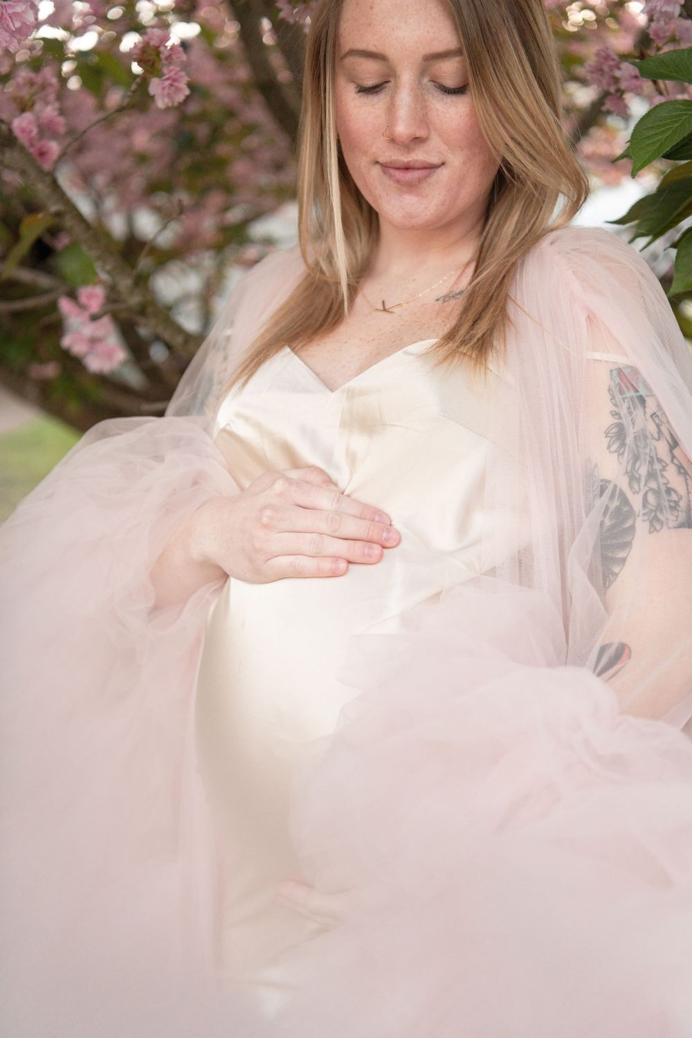 Expectant woman in a soft pink tulle dress poses among cherry blossoms in an ethereal maternity photo.