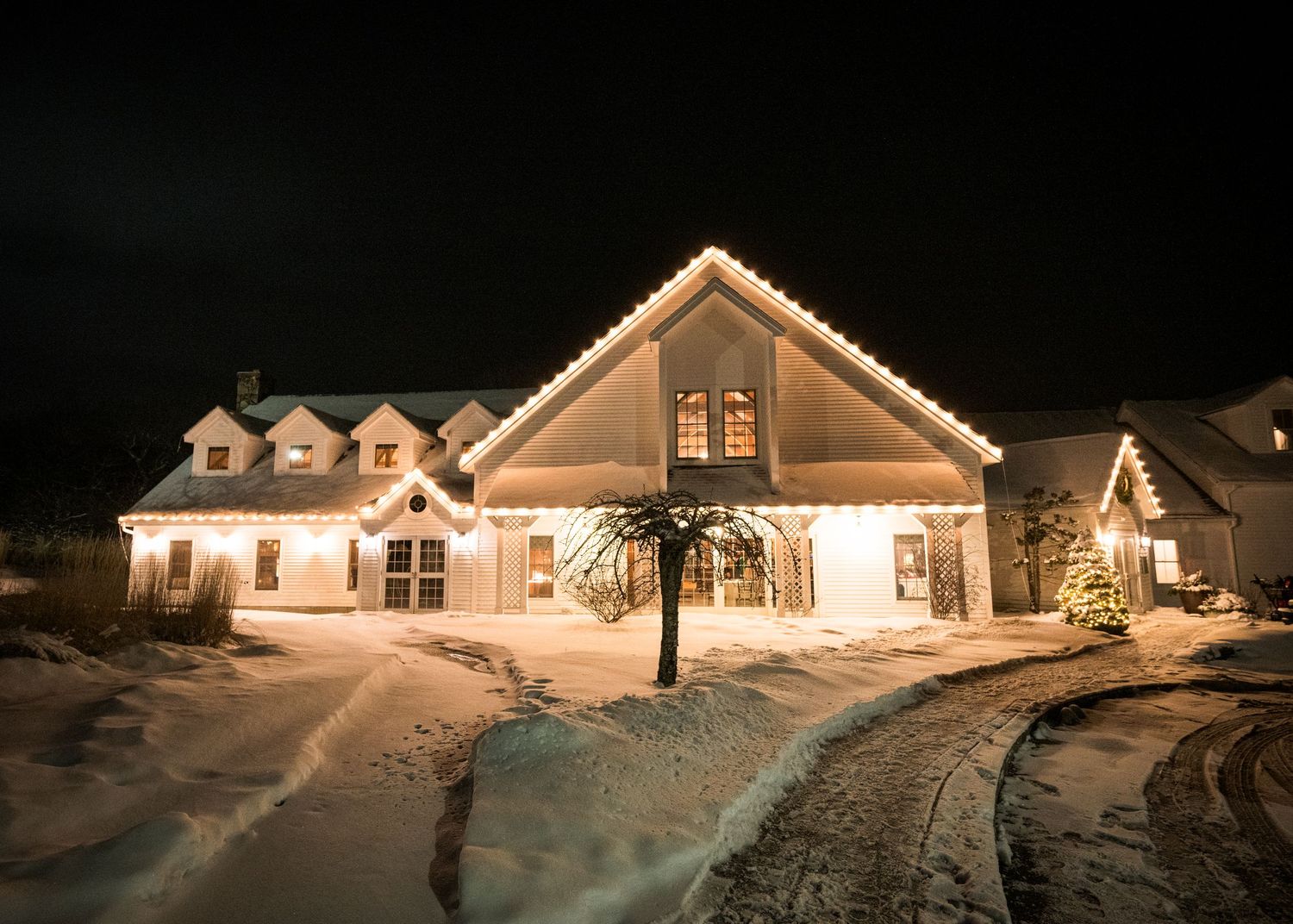 Large white house with dormers illuminated by warm Christmas lights in snowy winter landscape at night.