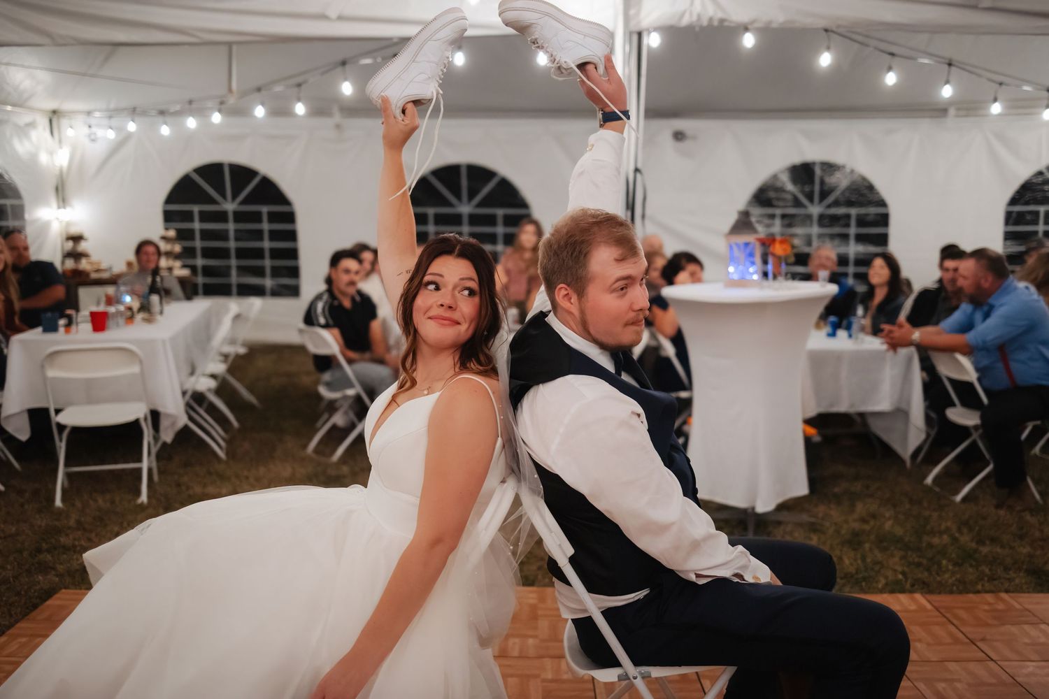 Newlyweds play a fun wedding reception game while seated back-to-back in a white tent venue with string lights.