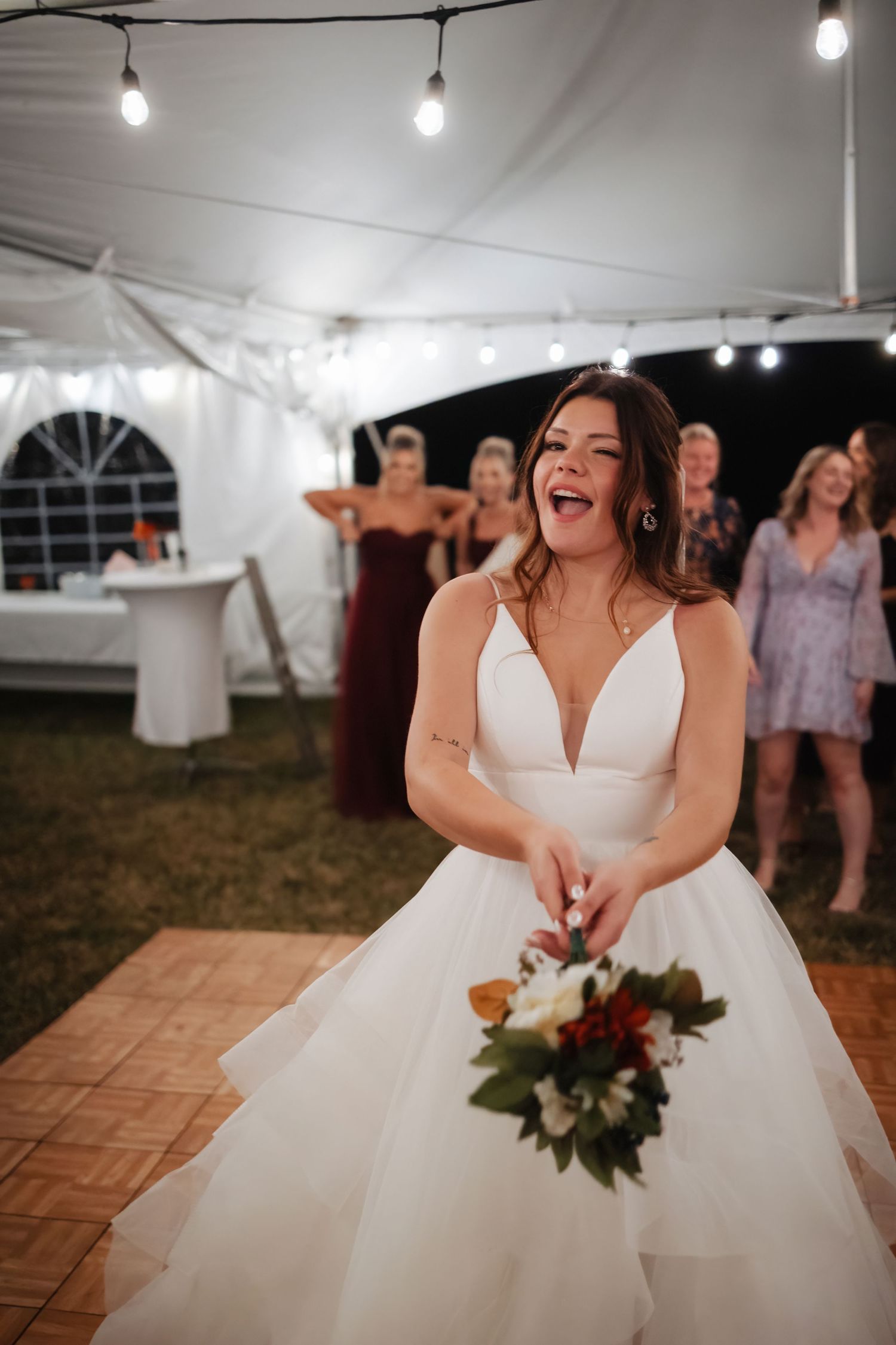 A joyful bride in a white ballgown holds her bouquet during the wedding reception under a tent with string lights.