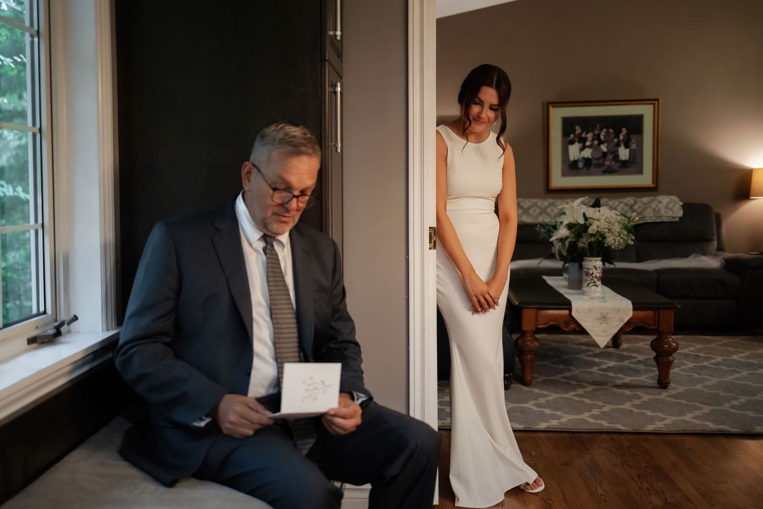An emotional moment captured in a living room as a bride in sleek white dress stands in doorway.